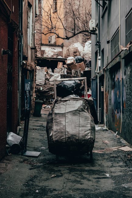 A collection of discarded wooden furniture and household debris piled against an outdoor brick wall, including a weathered wooden garden chair with slatted seat and backrest leaning on its side, a bro
